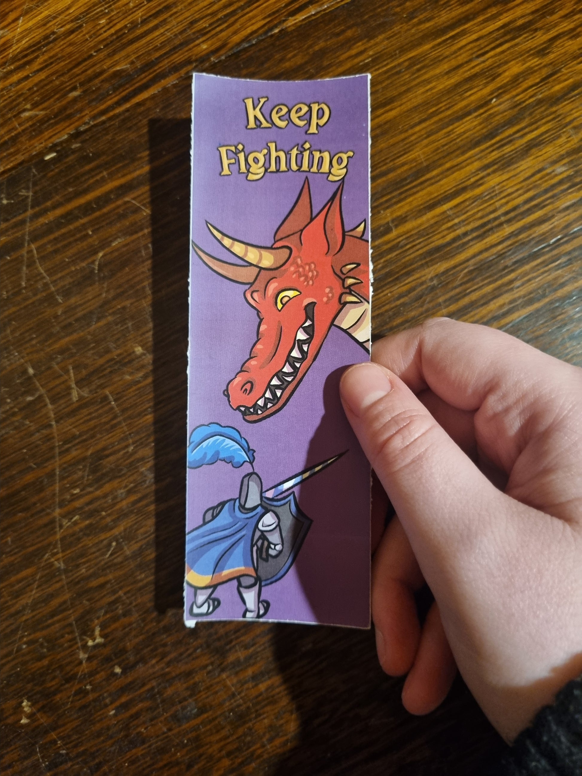 A hand holding a rectangular printed card or sticker featuring a purple background with a red dragon head, the words 'Keep Fighting' in yellow, and a cartoon knight in blue armor, placed on a wooden tabletop.