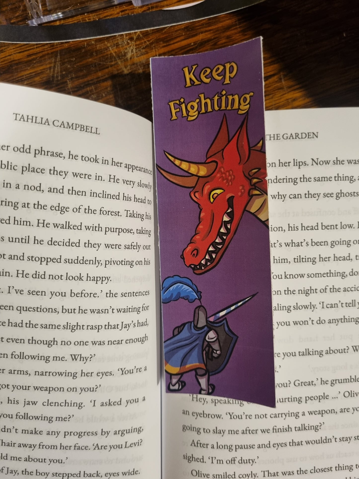 A purple book bookmark with a cartoon red dragon and a knight holding a sword, placed on an open book. The bookmark has the words 'Keep Fighting' printed on it.