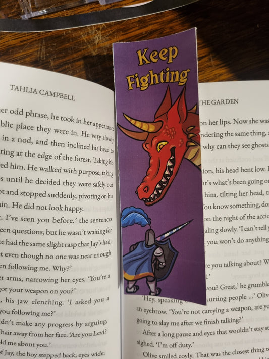 A purple book bookmark with a cartoon red dragon and a knight holding a sword, placed on an open book. The bookmark has the words 'Keep Fighting' printed on it.